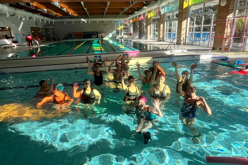 Un groupe d’aquagym dans le petit bassin, bras levés, sous le soleil qui traverse les vitraux.