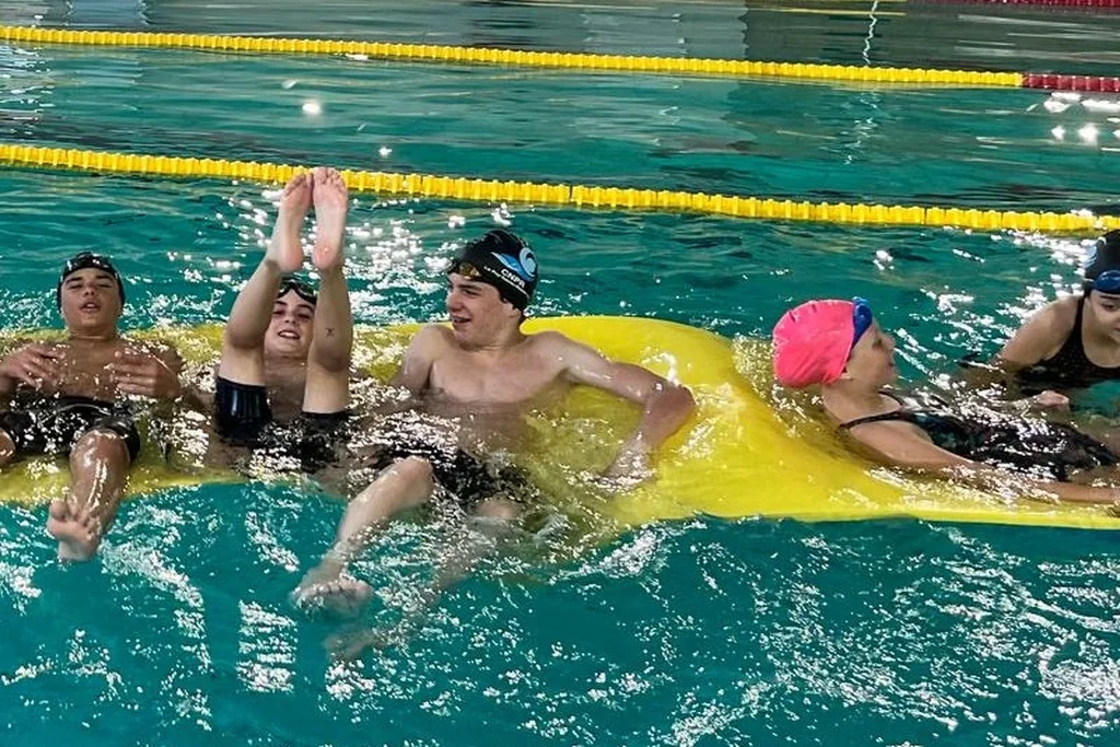 Des jeunes du club rient, allongés sur un tapis flottant jaune dans le bassin, un jour de fête.