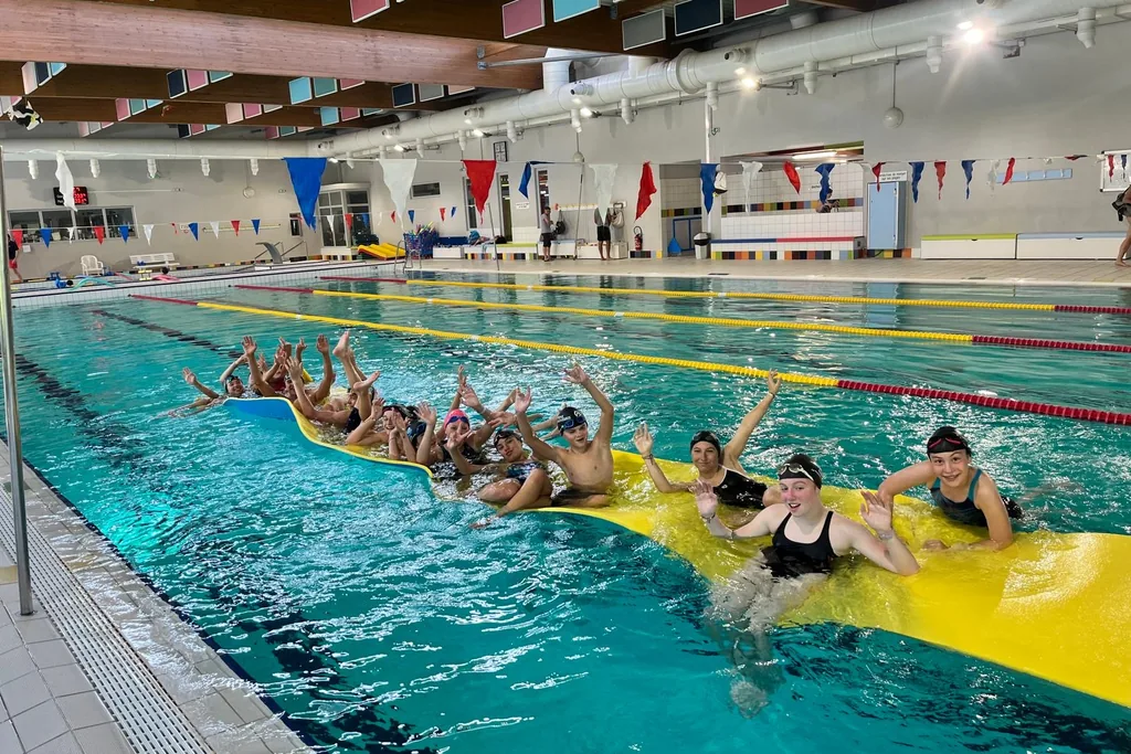 Des jeunes du club sur un grand tapis flottant jaune, dans le bassin, font coucou.